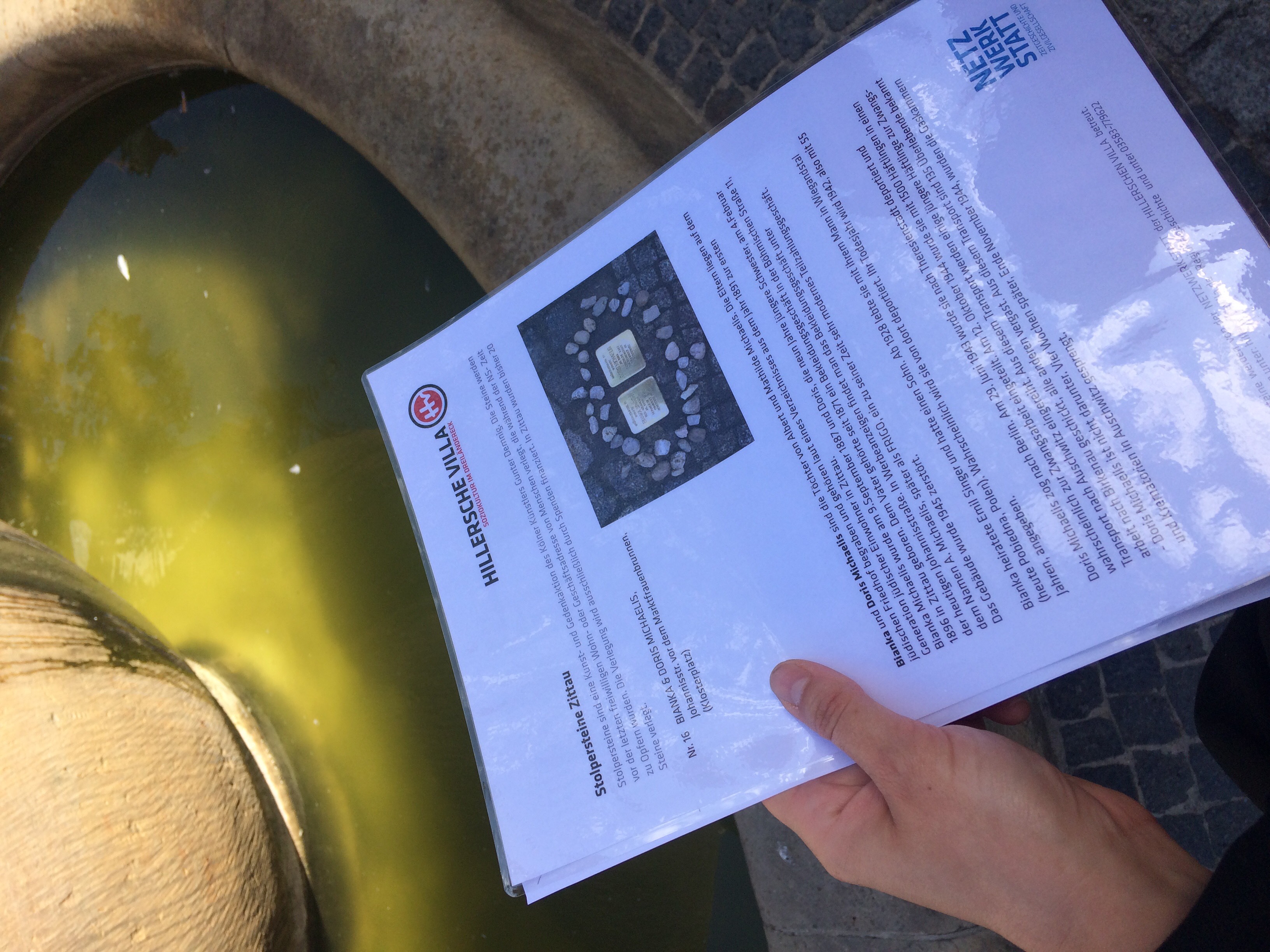 Das Bild zeigt eine Hand, die ein Handout hält, das Informationen über Stolpersteine in Zittau beinhaltet.
